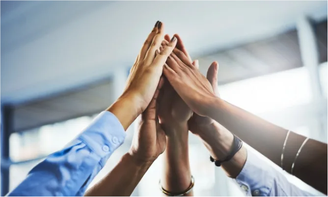 Six colleagues reaching up to meet in a high five in a bright office.